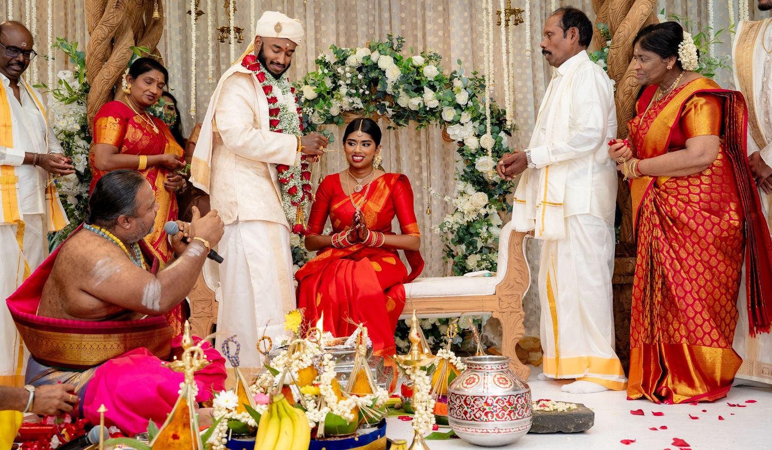 Hindu wedding ceremony with priest conducting rituals as bride in red and groom in white stand at mandap.