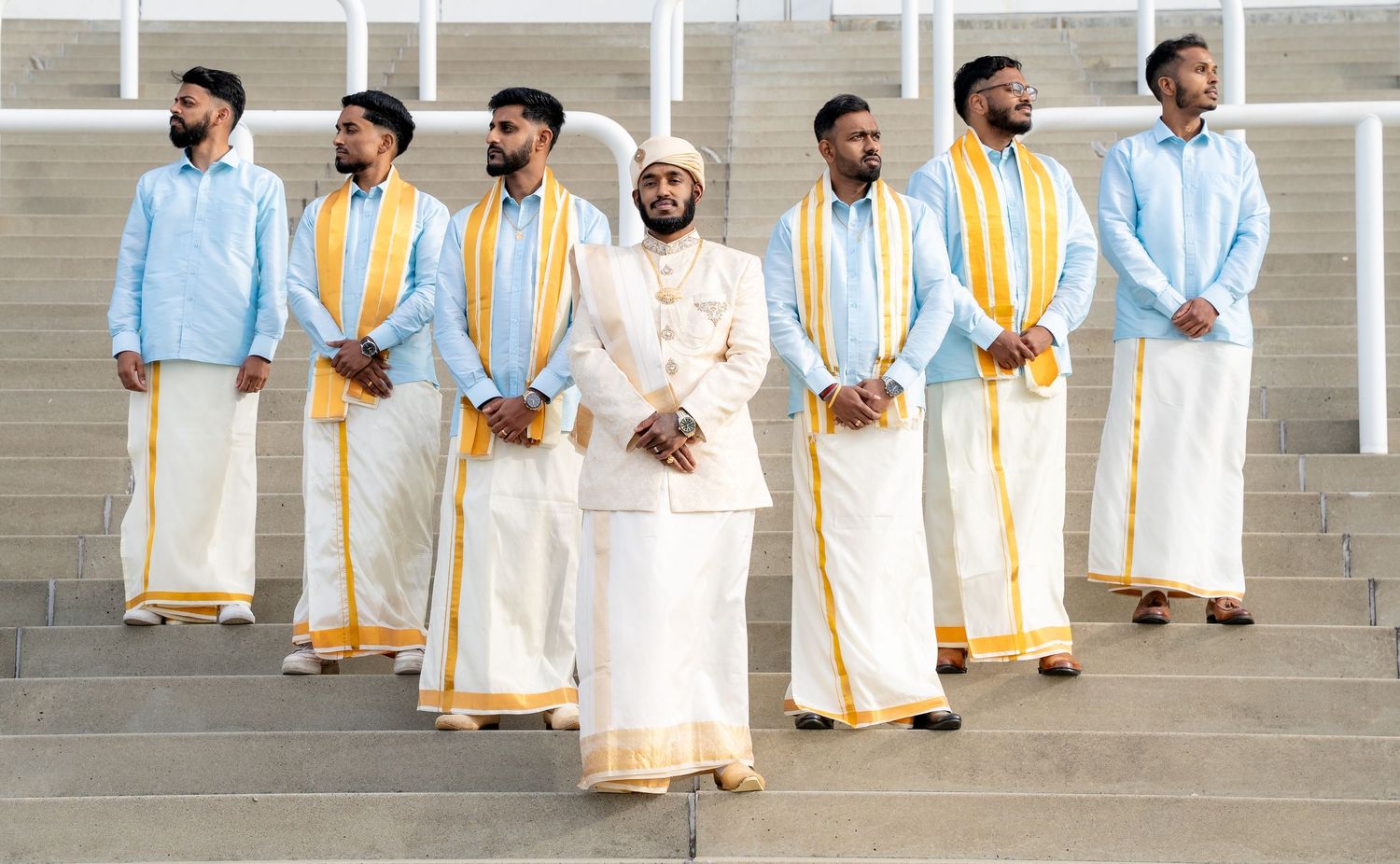 Groom in white and gold sherwani stands with groomsmen wearing light blue shirts and white dhotis on steps.