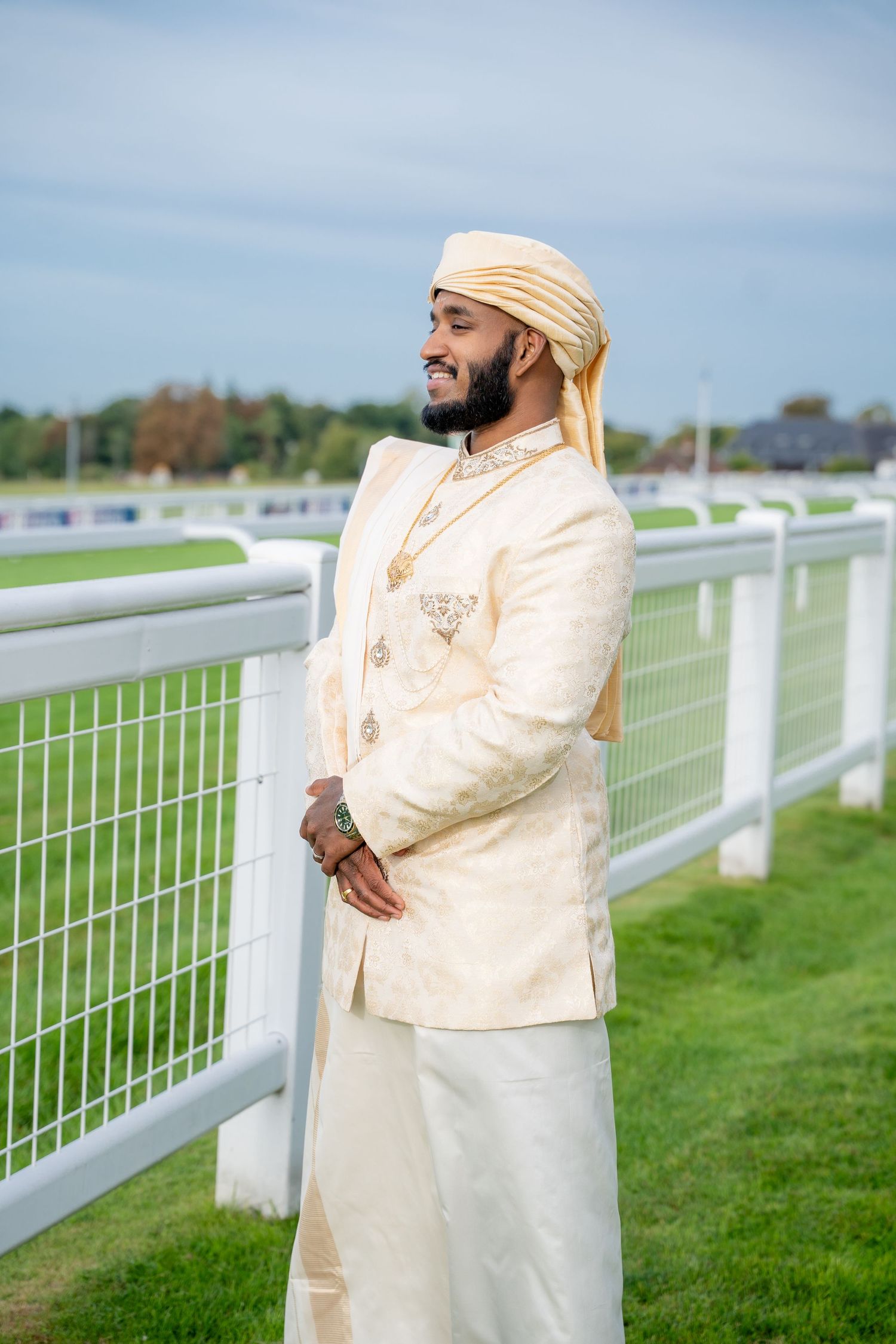 A smiling man in traditional Indian sherwani and turban stands by a white fence on a sunny day.