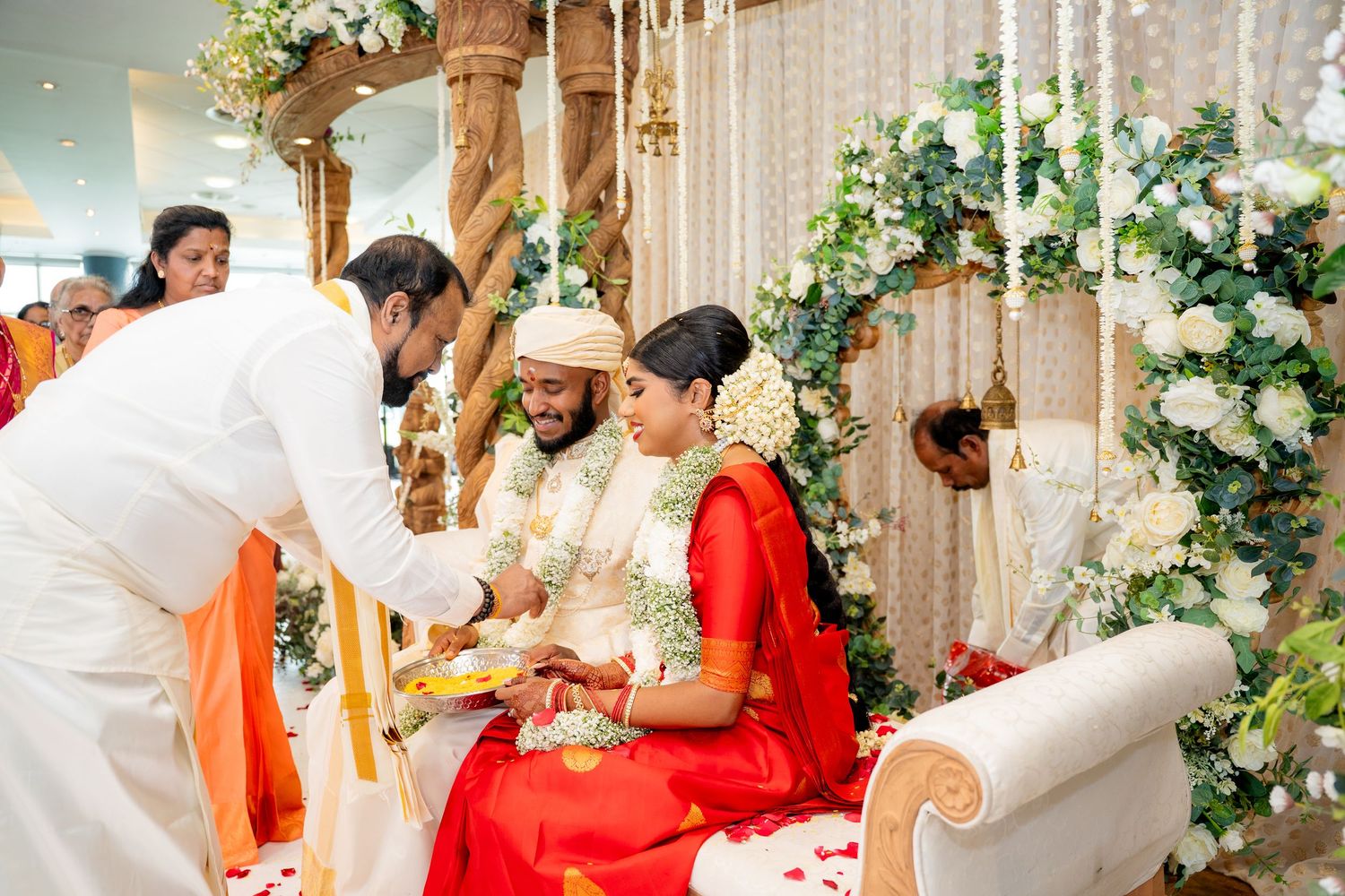 Indian bride and groom in traditional red and gold attire during Hindu wedding ceremony with floral decorations.