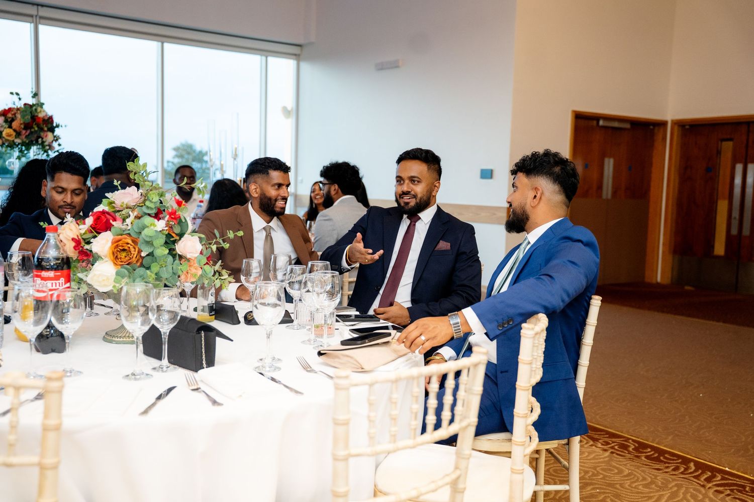 Men in suits laughing and conversing at an elegantly decorated wedding reception dinner table.