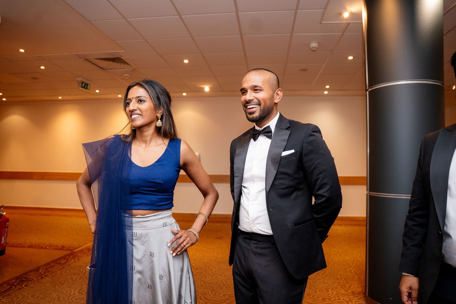Elegant couple in formal attire, woman in blue gown and man in black tuxedo, smiling at an indoor event.