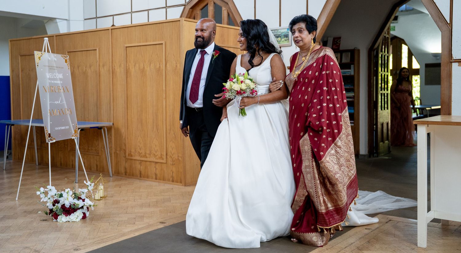 Bride in white gown walks with groom in red tie and woman in red sari at church wedding ceremony.