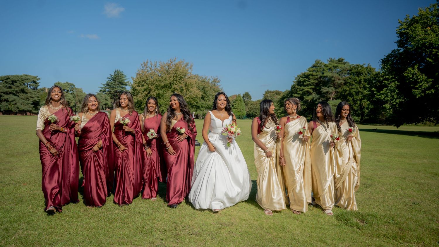 Bride in white ball gown poses with bridesmaids in burgundy and gold sarees on sunny lawn.