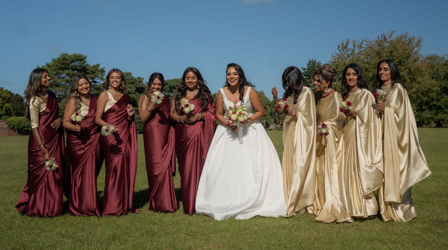 Bride in white gown poses with bridesmaids in burgundy and gold saris on sunny green lawn.