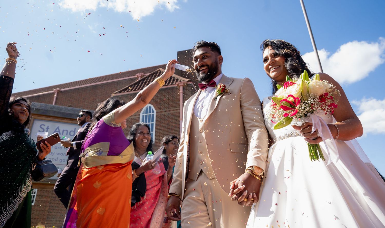 Joyful newlywed couple holding hands outside church as guests throw confetti during vibrant multicultural wedding celebration.