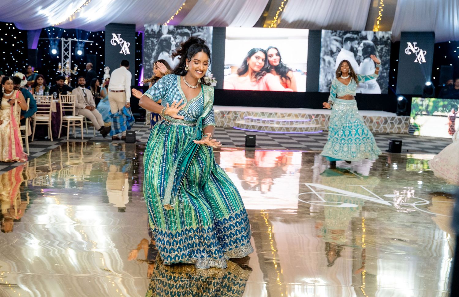 Woman dancing in blue maxi dress on dance floor at Indian wedding reception with screen backdrop.