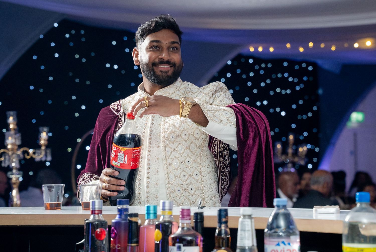 Man in traditional Indian sherwani smiles while holding a Coca-Cola bottle at a wedding reception bar.