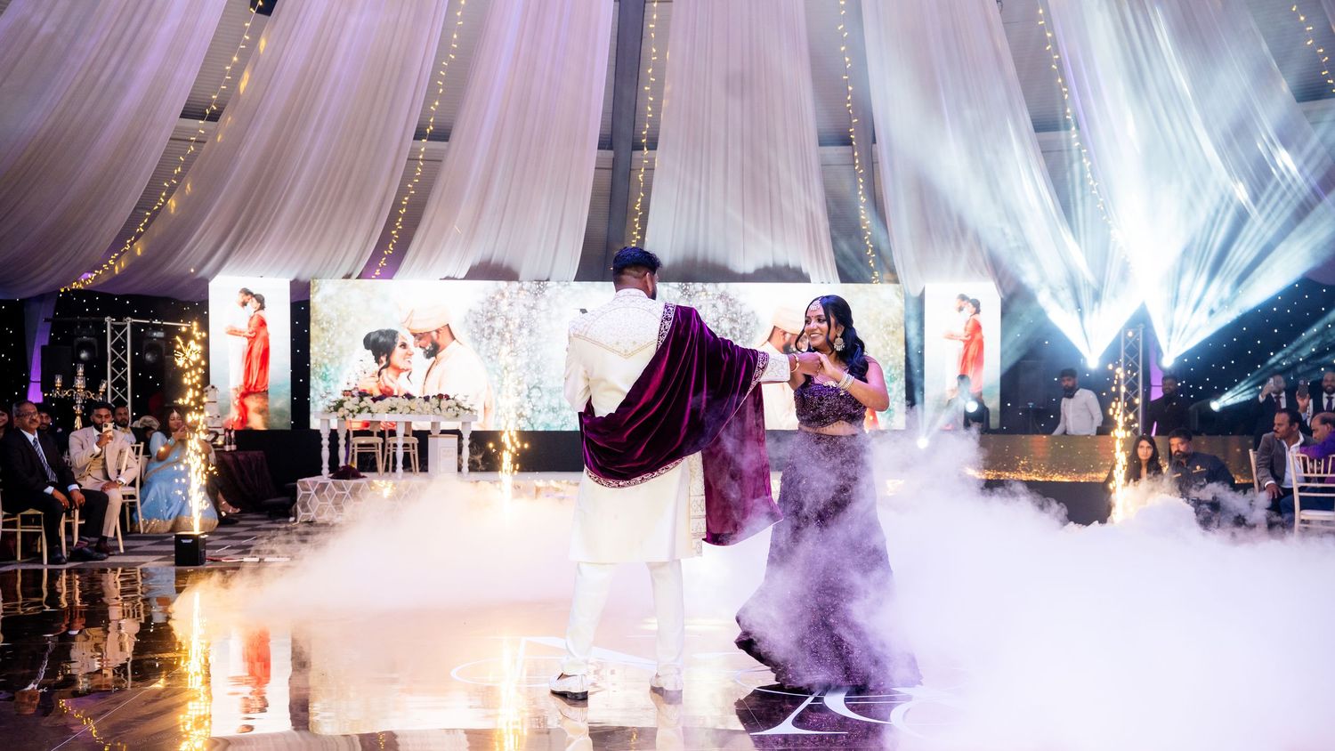 Indian couple sharing first dance surrounded by dramatic fog effects on illuminated wedding reception stage.
