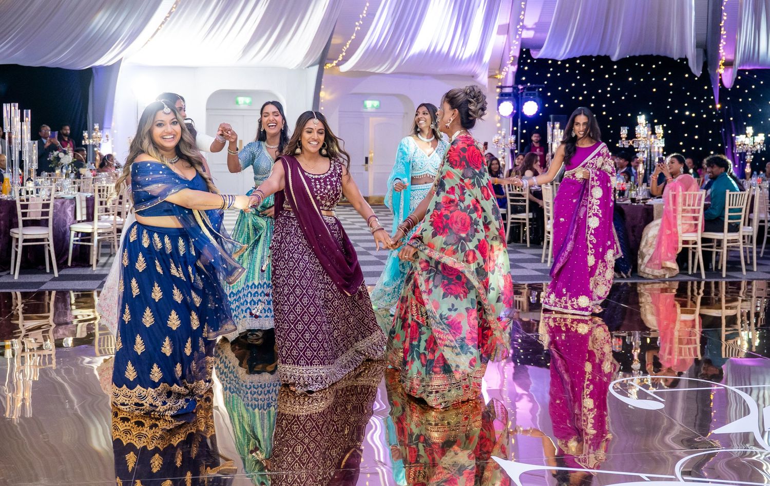 Women in traditional Indian wedding outfits dancing joyfully on a mirrored reception dance floor.