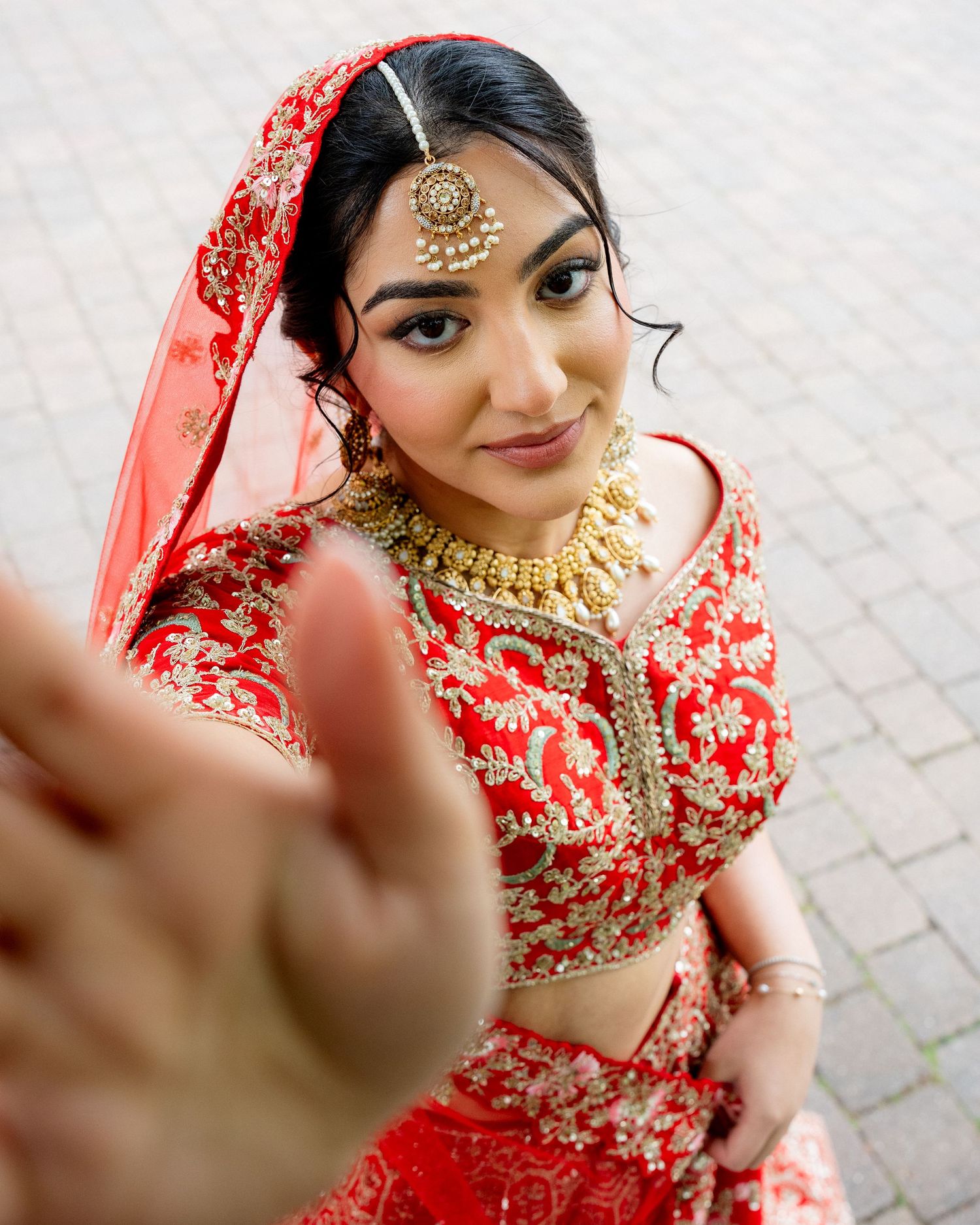 Indian bride in red and gold lehenga with maang tikka and gold jewelry reaches toward camera.