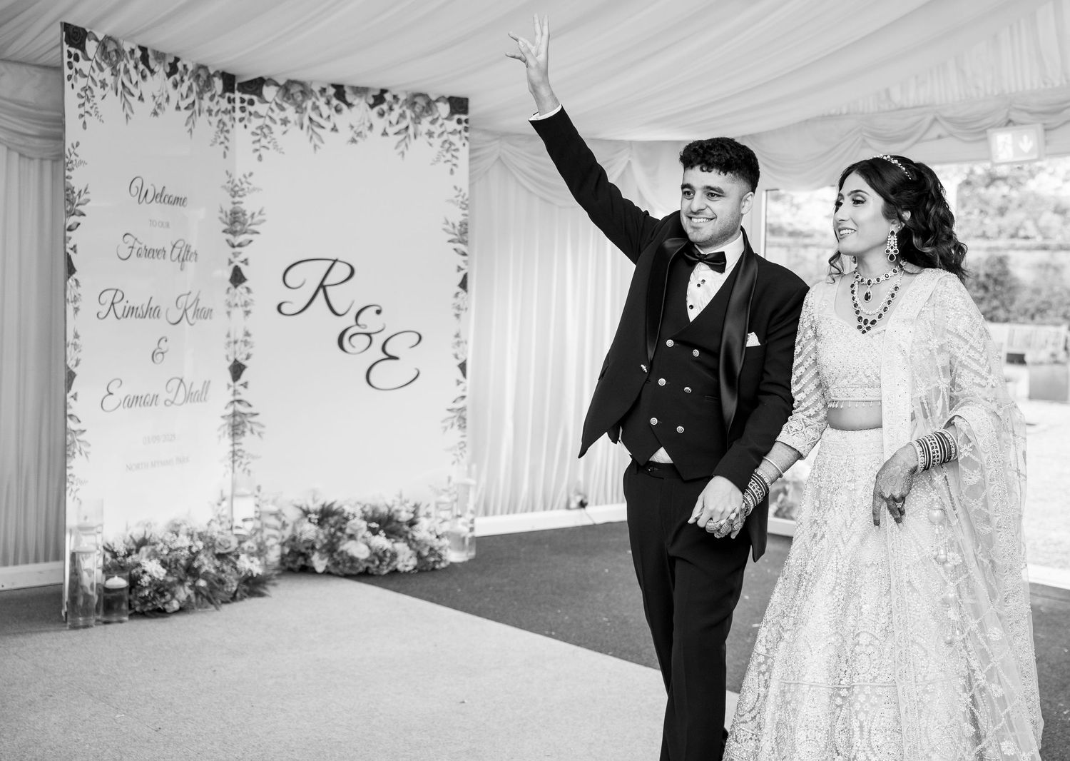 Black and white photo of a couple making a grand entrance at their wedding reception, posing in front of a floral monogram backdrop.
