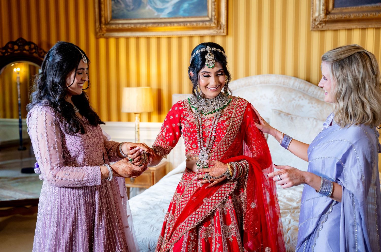Indian bride in red lehenga assisted by two women during wedding preparation in ornate room.