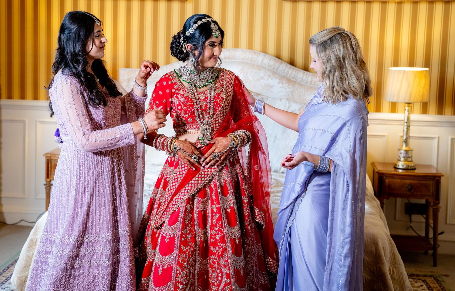 Bride in red bridal lehenga being dressed by two women in a elegant bedroom setting.