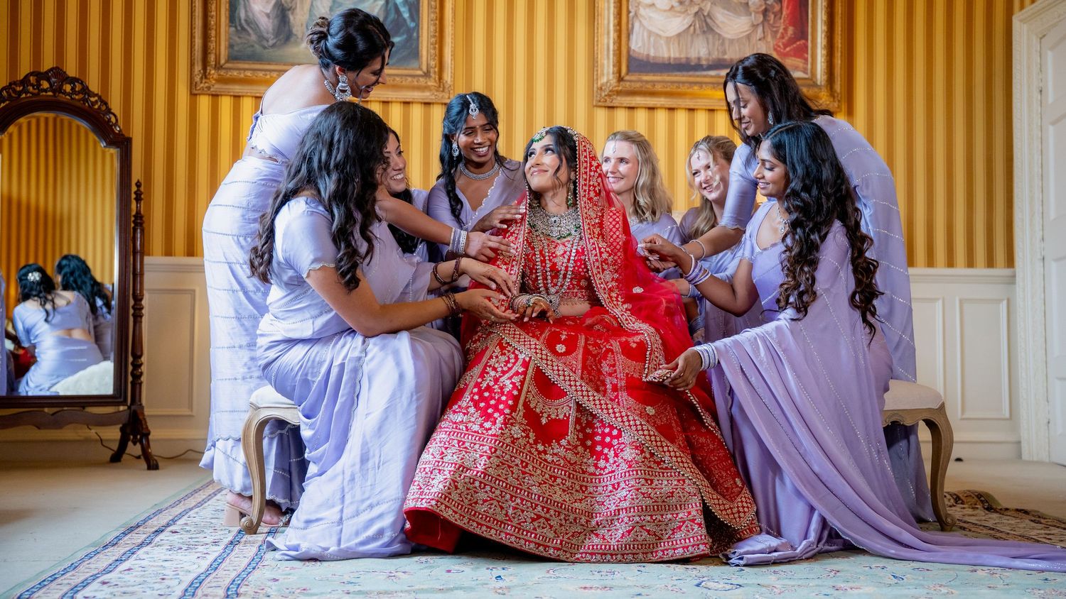 Indian bride in red lehenga surrounded by bridesmaids in lavender sarees during bridal preparation ceremony.