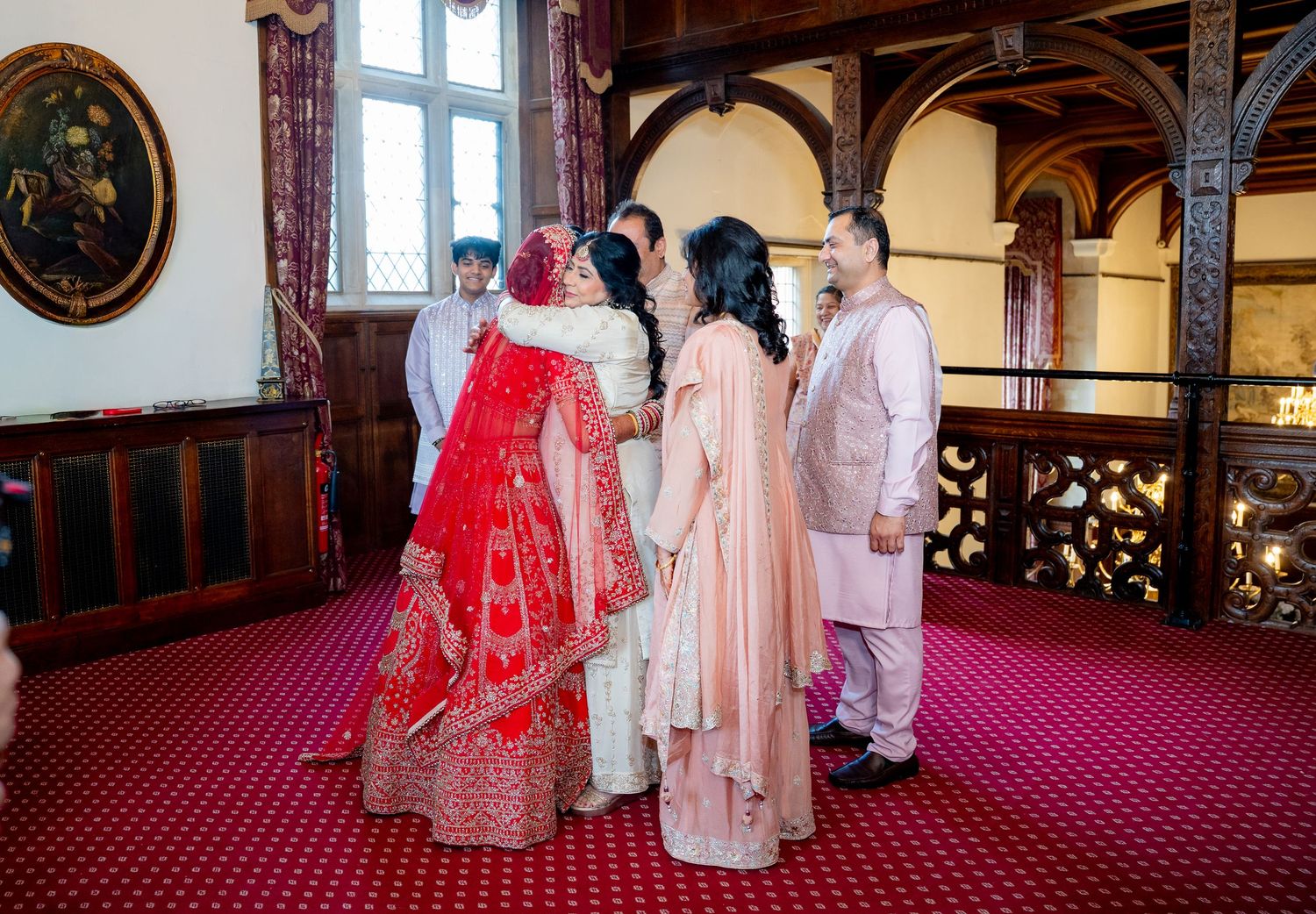 Indian bride in red lehenga shares emotional embrace with family at traditional wedding.