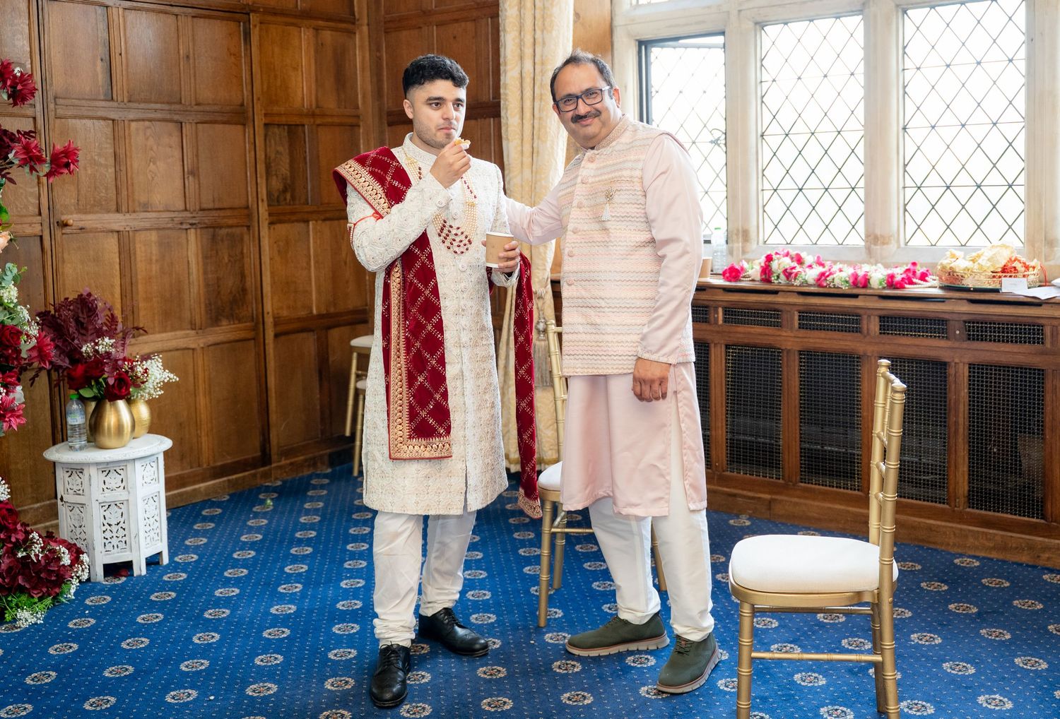 Groom in white and red sherwani stands with an elder man in a blue-carpeted wedding venue room.