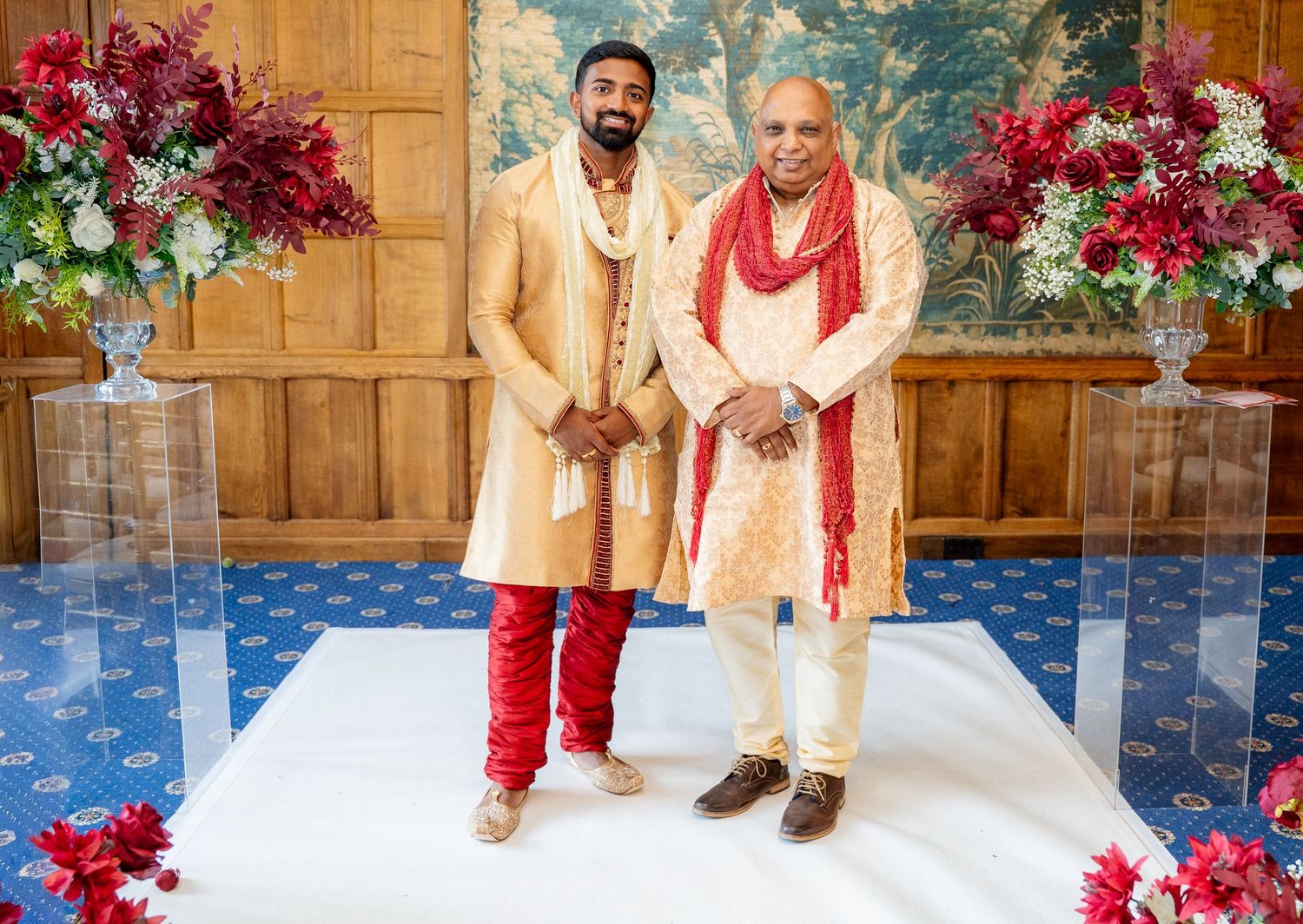 Two men in traditional Indian wedding attire stand together at an elegant venue with floral arrangements.