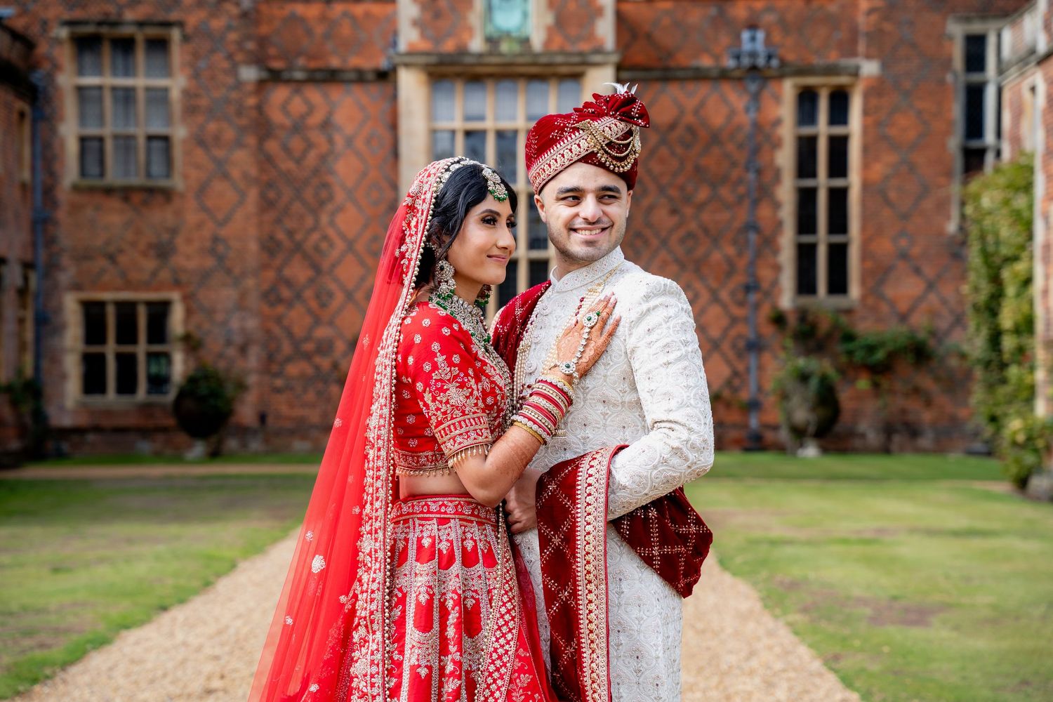 Indian bride and groom in matching red and white traditional wedding attire posing lovingly at a brick manor.