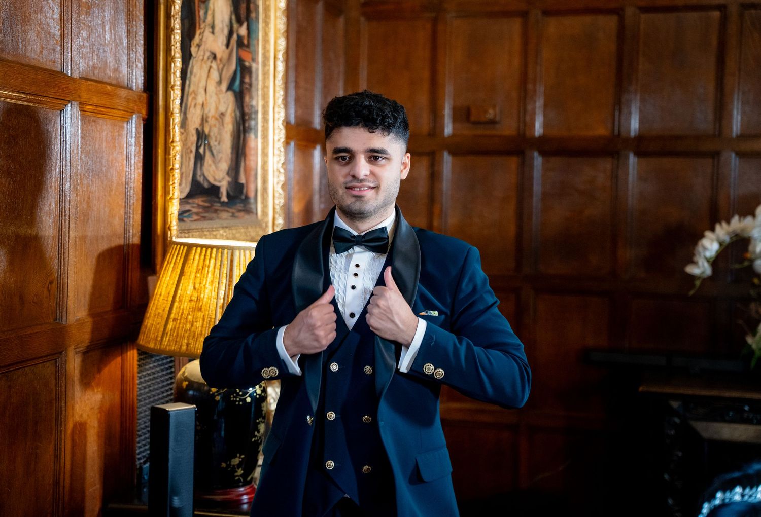 Young man in navy blue tuxedo with bow tie posing confidently in an elegant wood-paneled room.
