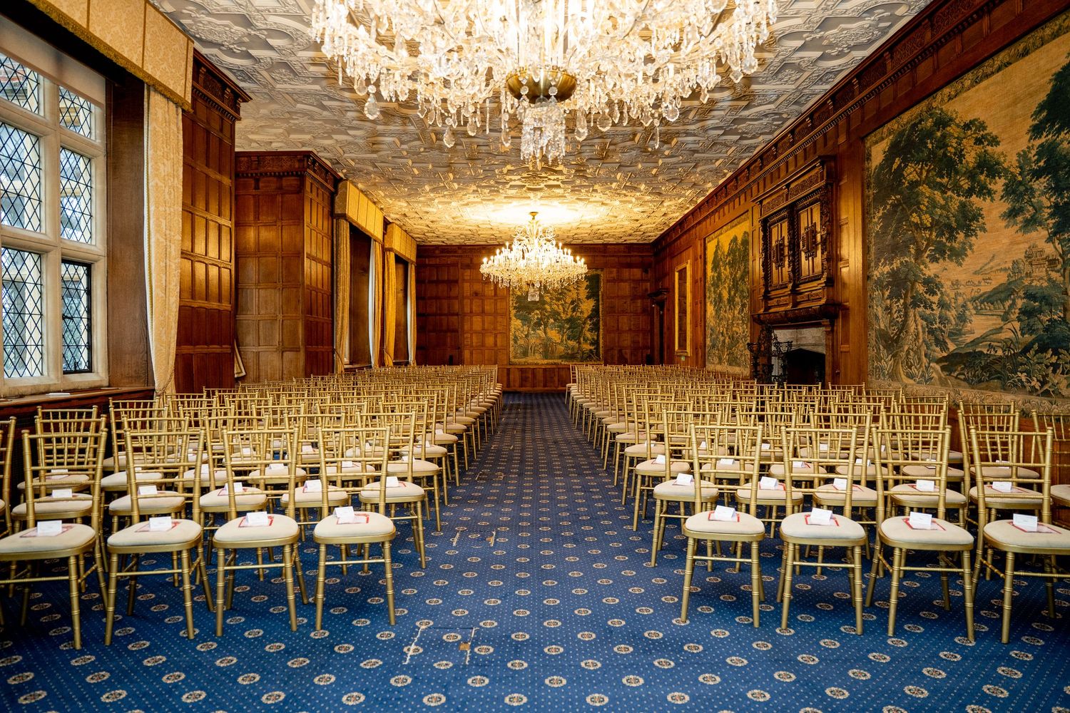 Elegant ballroom with gold Chiavari chairs, crystal chandeliers, blue patterned carpet, and ornate tapestry walls set for an event.
