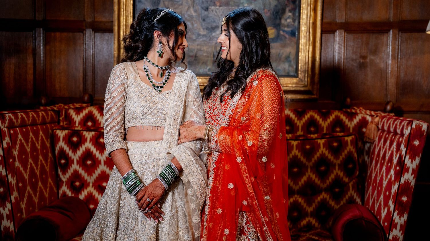 Two brides in stunning Indian bridal attire, one in ivory lehenga and one in red, share a romantic moment at their wedding.