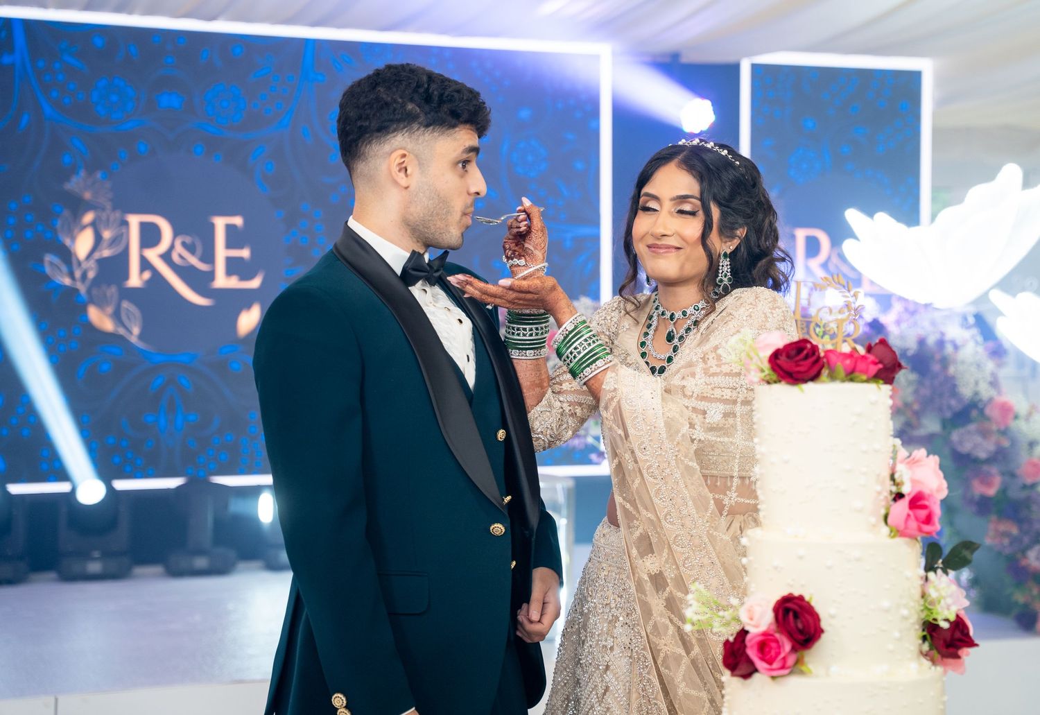 South Asian couple in elegant wedding attire cutting a white tiered cake at a reception venue.