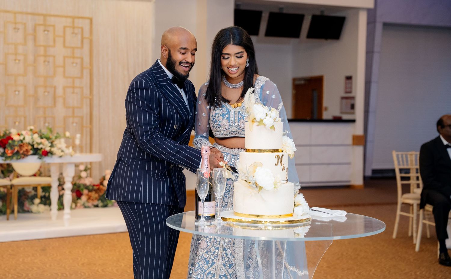 Smiling couple in wedding attire cuts white tiered cake at decorated reception venue.