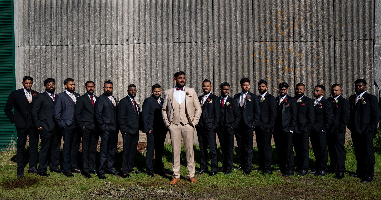 Groom in tan suit stands centered among groomsmen in dark suits before a metal wall.