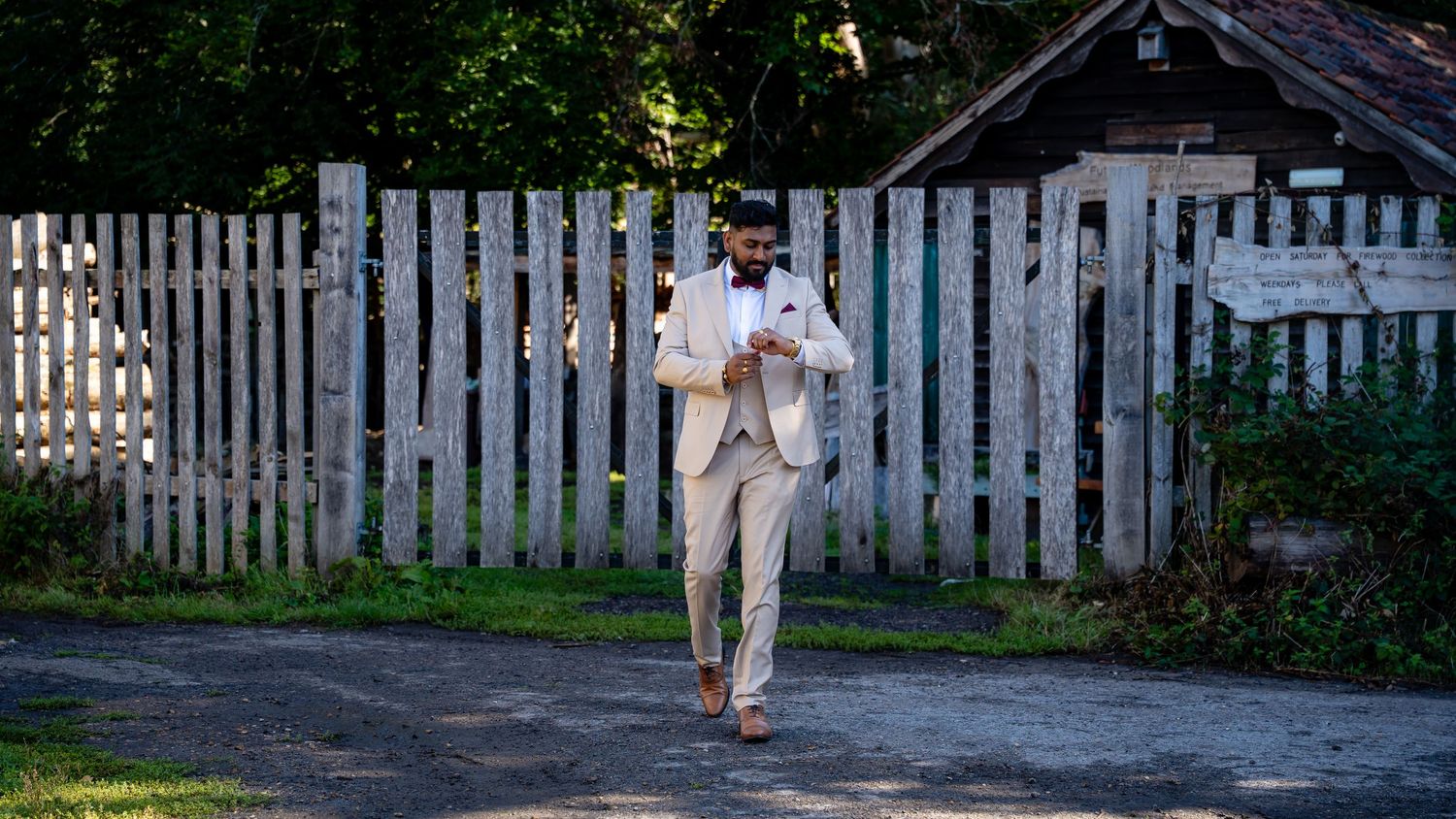 Groom in cream suit checks phone while walking along a sunny rural pathway.