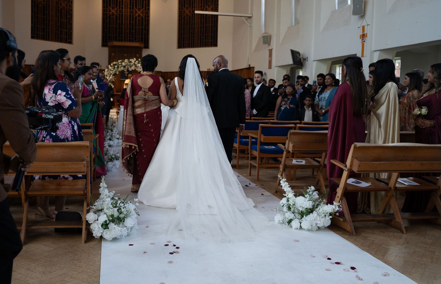 Bride in white gown and veil walks down church aisle with floral arrangements during wedding ceremony.