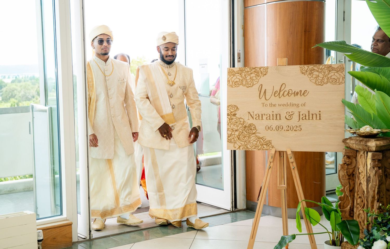 Two grooms in traditional white and gold Indian wedding outfits enter beside a wooden welcome sign.