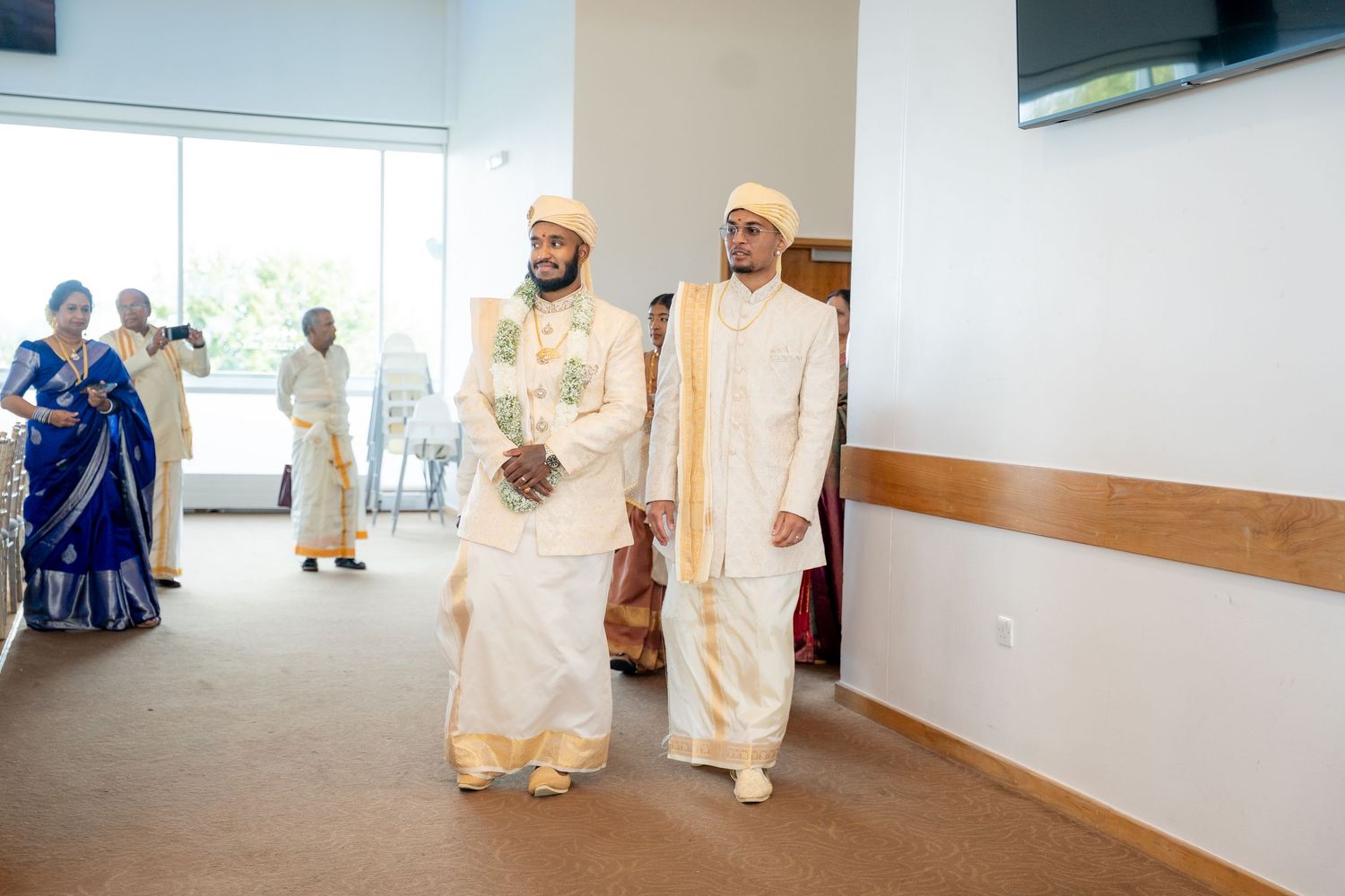 Two men in traditional white and gold South Asian wedding attire walking together at a formal ceremony.