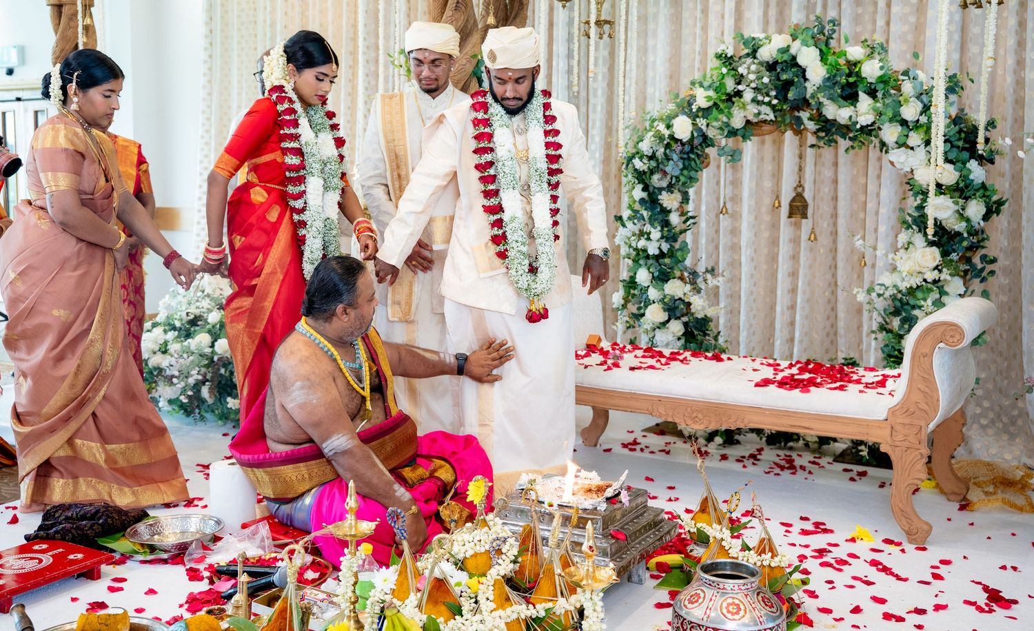 South Indian Hindu wedding ceremony with priest performing rituals as bride in red saree and groom in white stand on mandap.