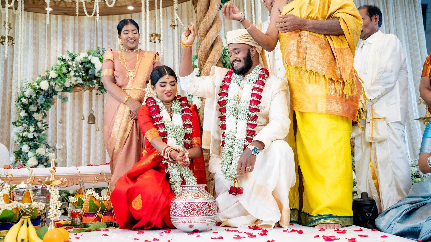 Bride in red saree and groom in white sitting at flower-adorned altar during Hindu wedding.