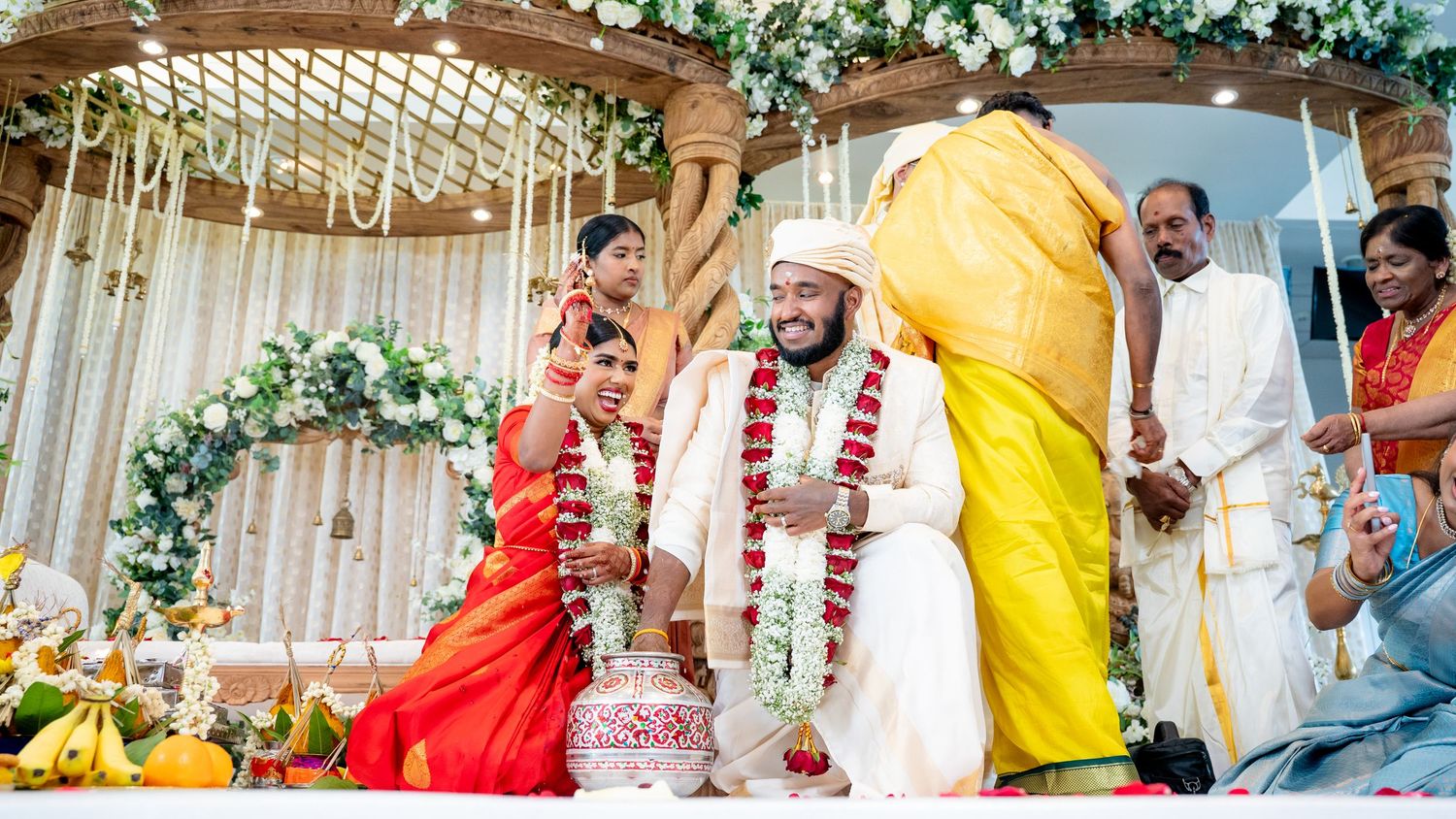Smiling groom in white and bride in red saree participate in Hindu wedding rituals on a decorated stage.