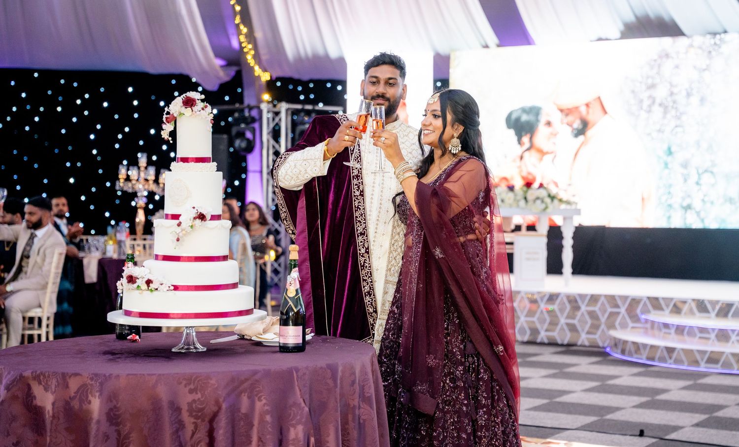 South Asian couple in burgundy and gold wedding outfits celebrating beside a decorated white wedding cake.