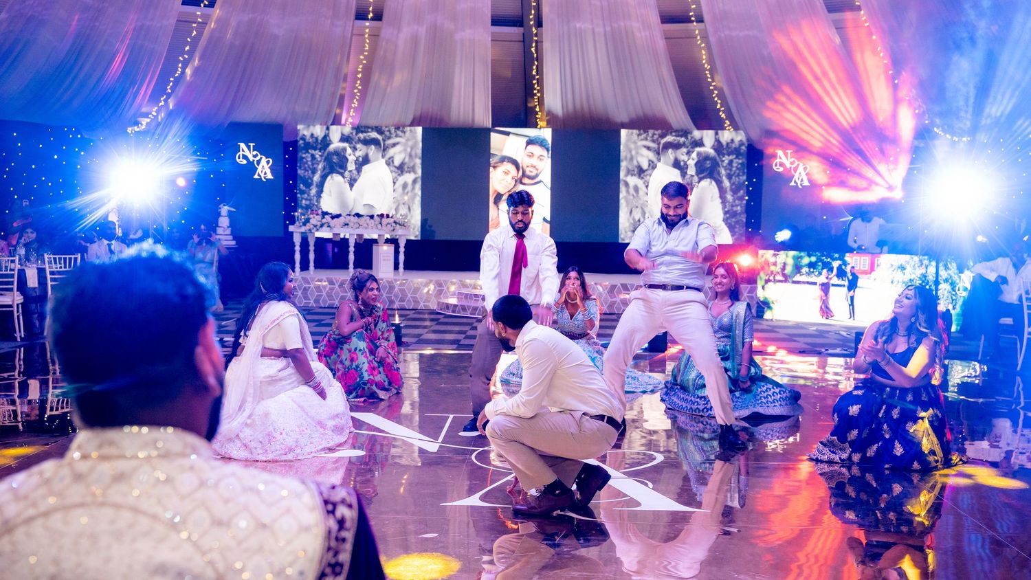 Colorful Indian wedding reception with guests dancing on stage under vibrant purple and blue lights at an elegant venue.