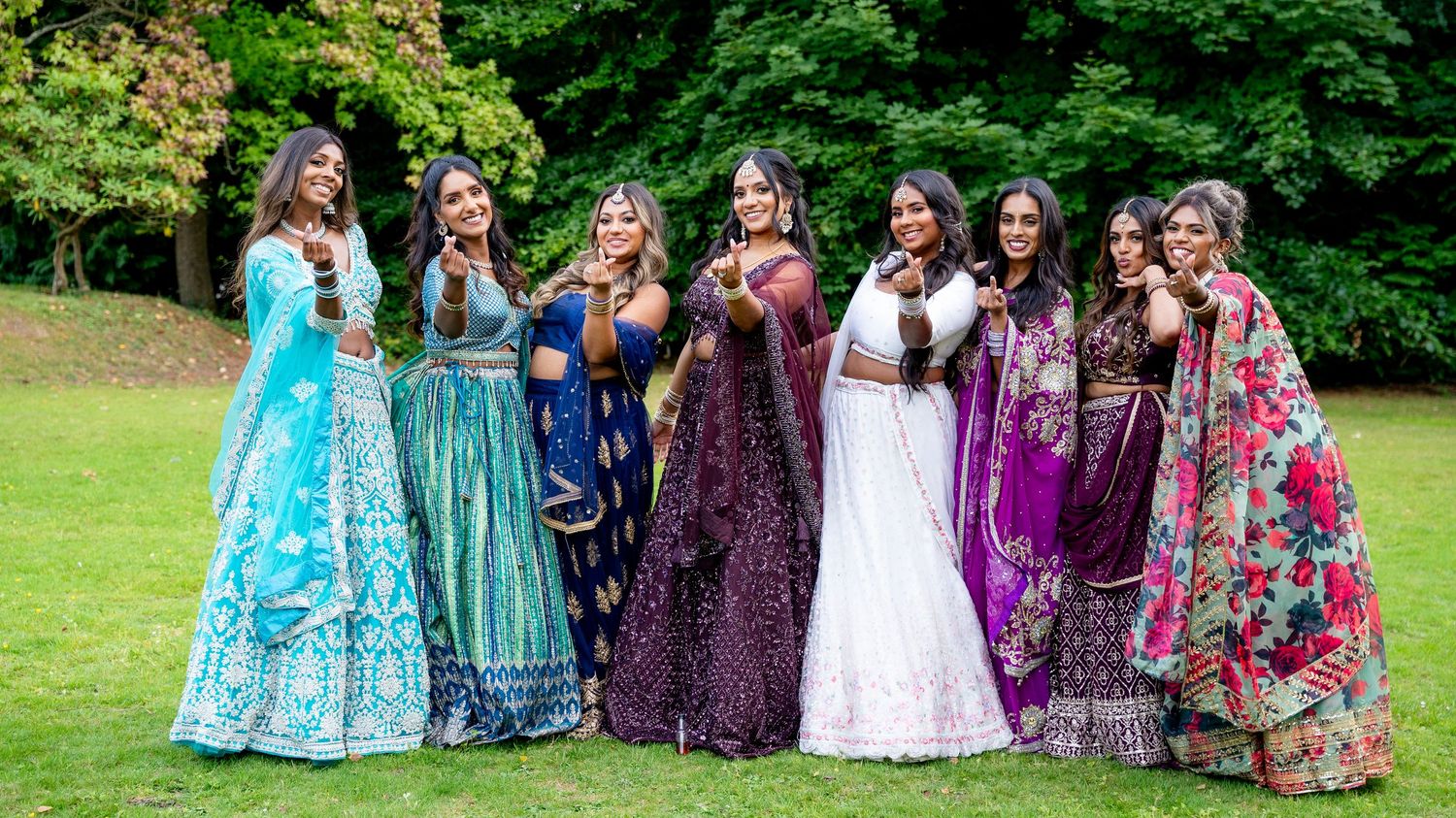 Seven women in vibrant traditional Indian lehenga outfits posing together outdoors on green grass with lush trees behind them.