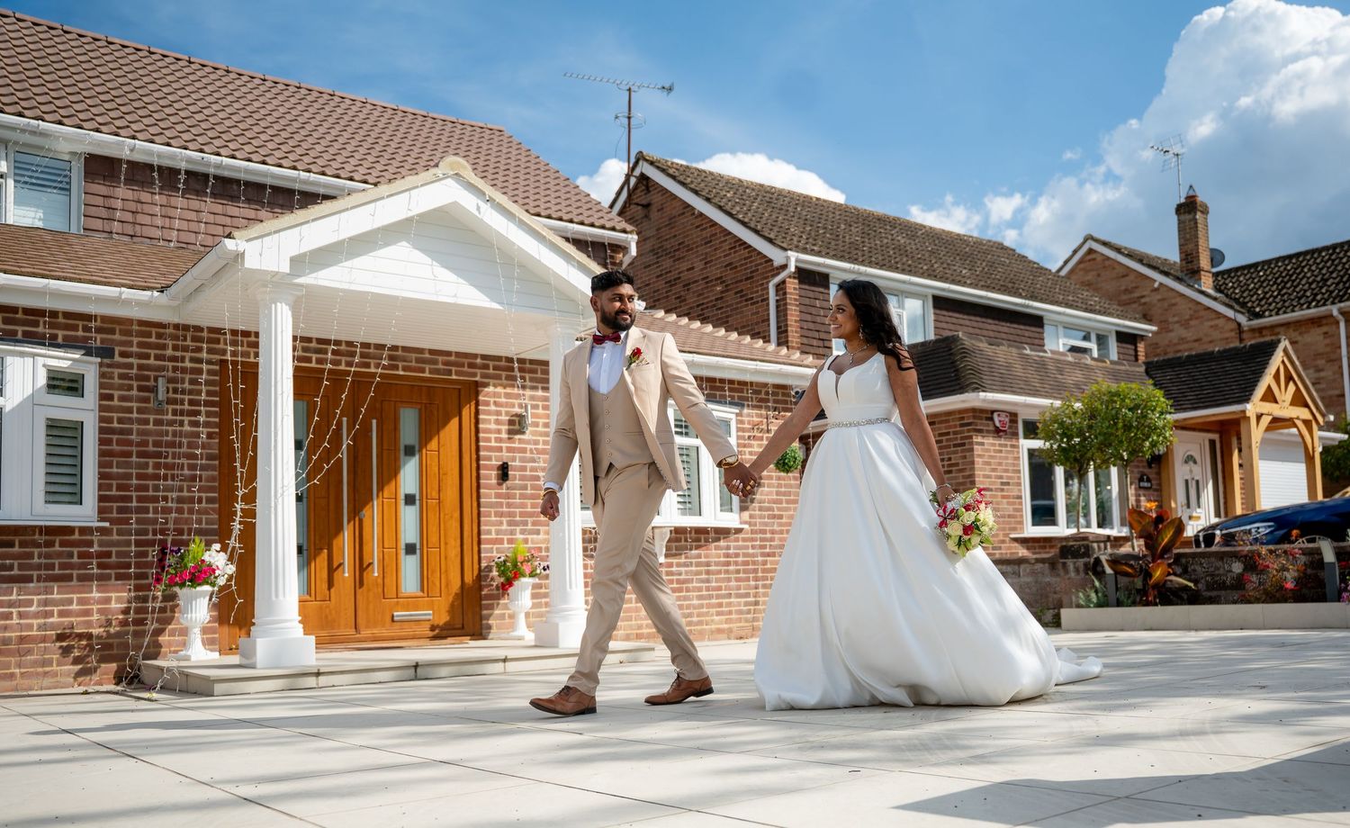 Bride and groom walking hand in hand outside a brick house on their wedding day, wearing elegant white gown and beige suit.