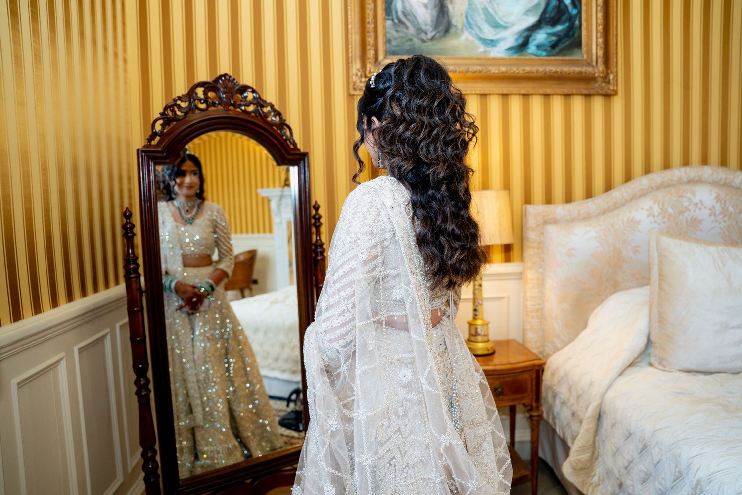 Bride in white embroidered lehenga admires her reflection in ornate mirror in elegant gold-wallpapered bridal suite.