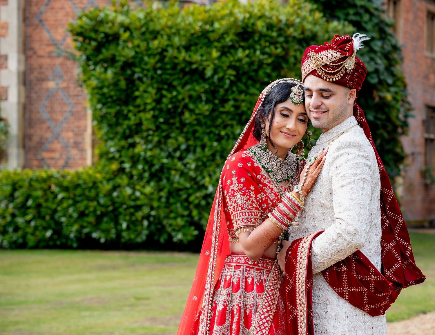 Indian bride in red lehenga and groom in white sherwani pose together outdoors at a traditional South Asian wedding.