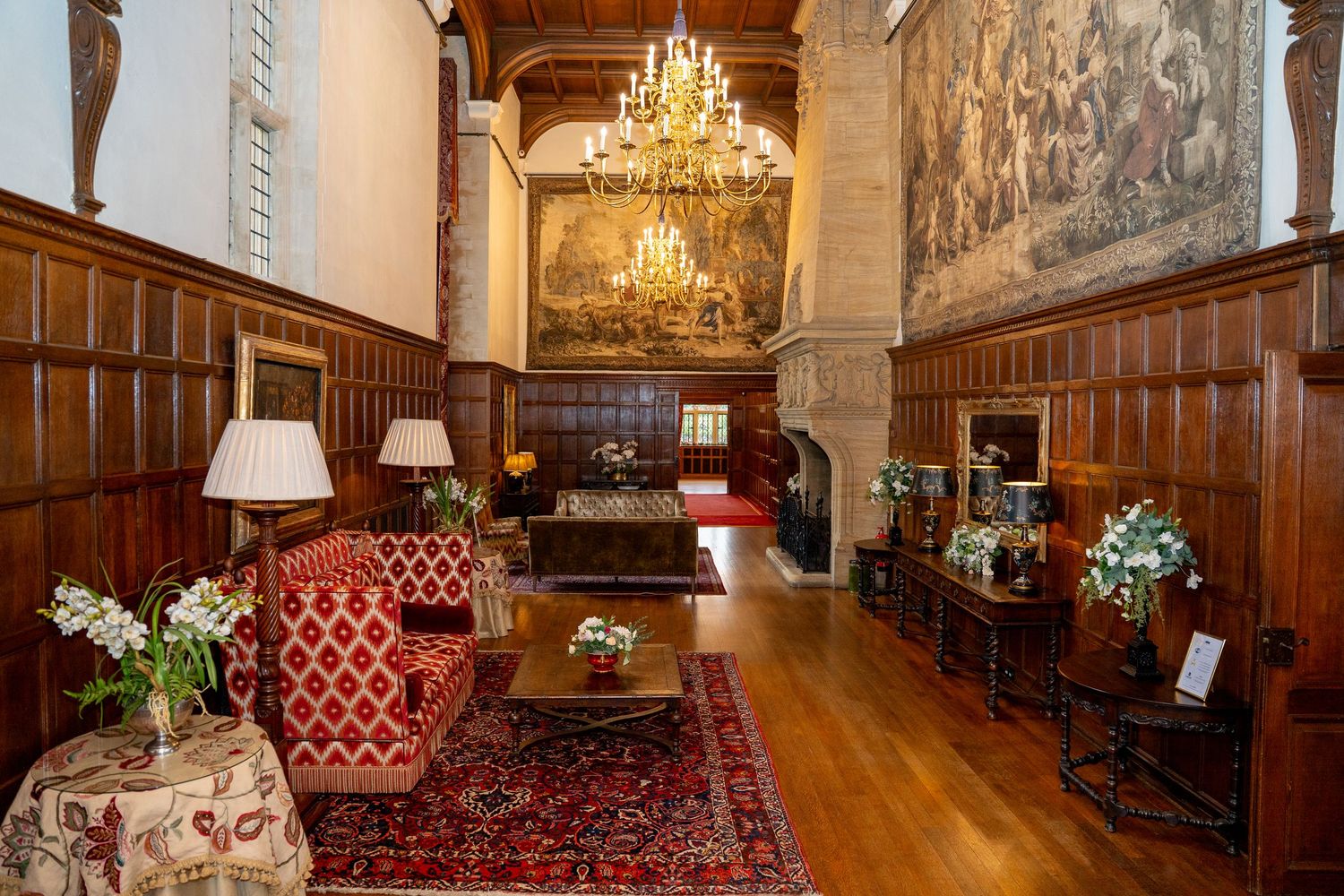 Grand historic manor hall with ornate chandelier, tapestry, red patterned sofas and Persian rug.