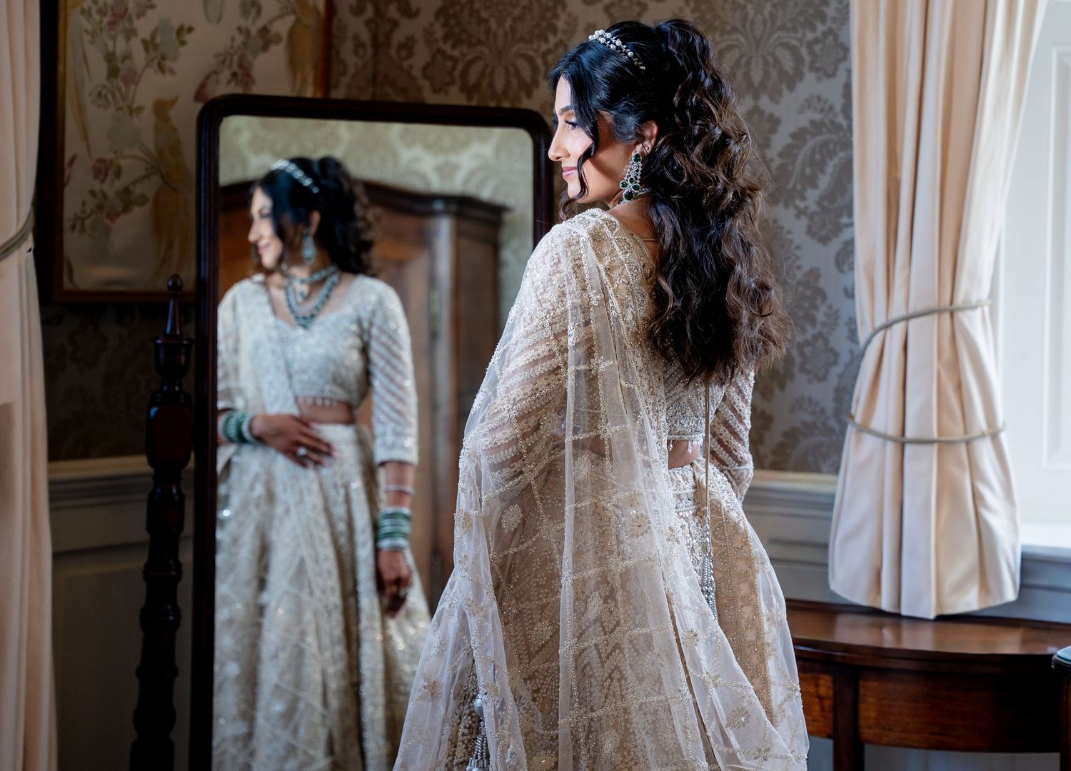 Bride in white embroidered lehenga admires her reflection in a full-length mirror in an elegant room.