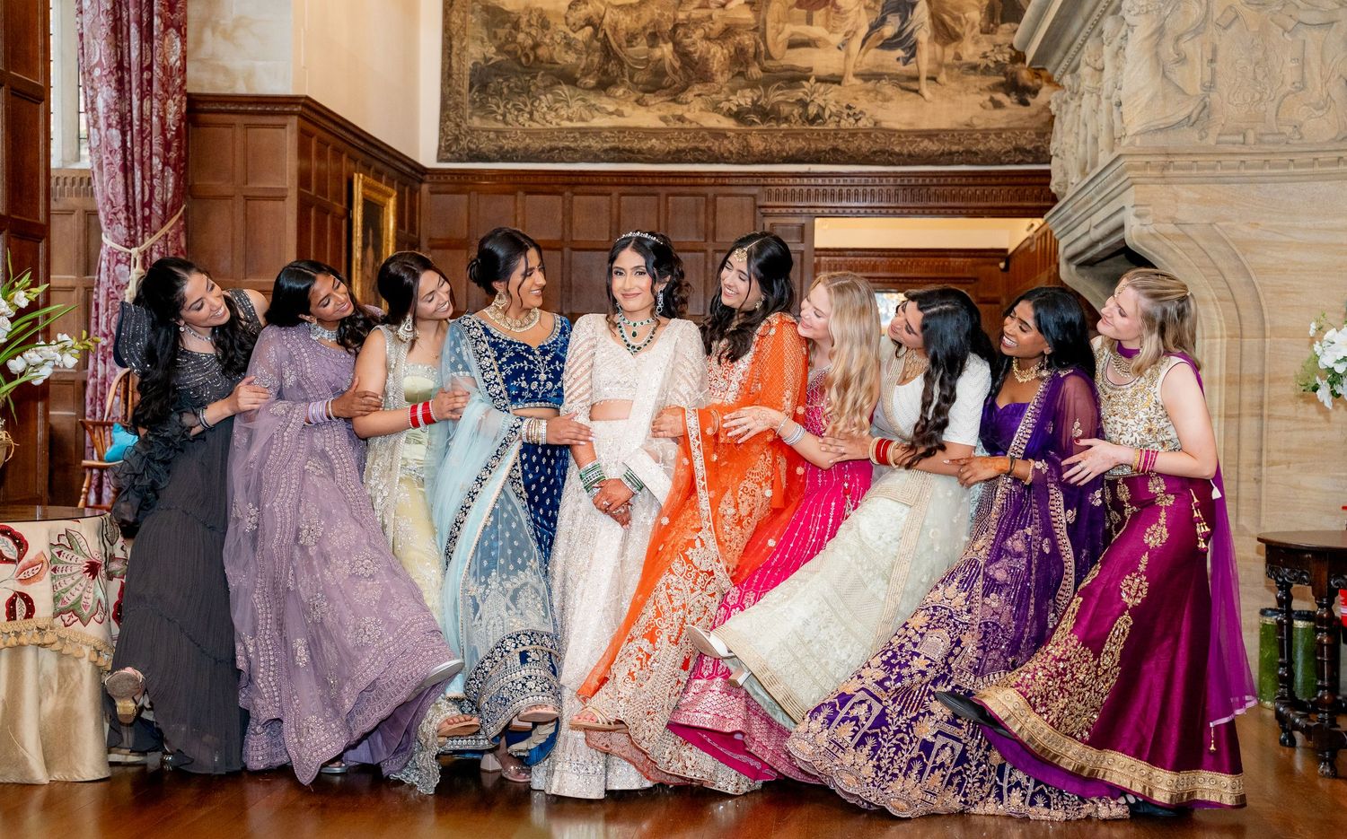 Bridal party in vibrant Indian lehenga outfits laughing together in an elegant ballroom setting.