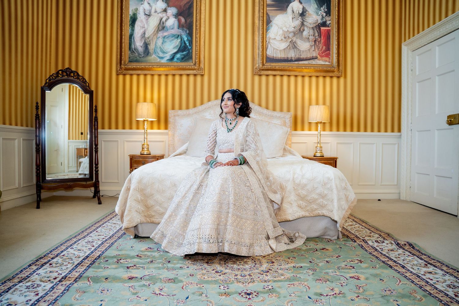 Bride in ivory embroidered lehenga sits elegantly on bed in ornate yellow striped room with antique mirror and paintings.