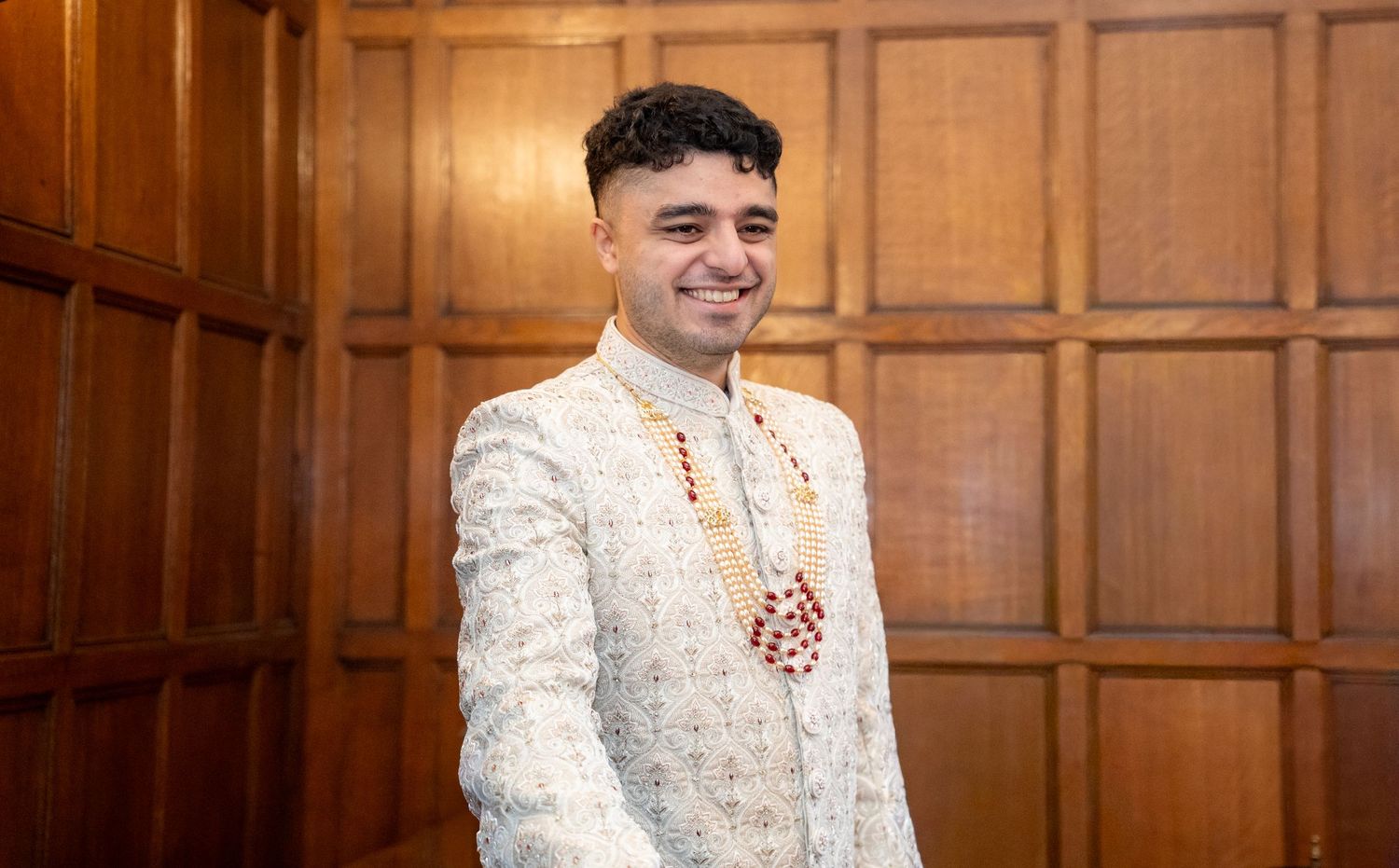 Smiling groom in embroidered sherwani with floral garland against wooden paneling.