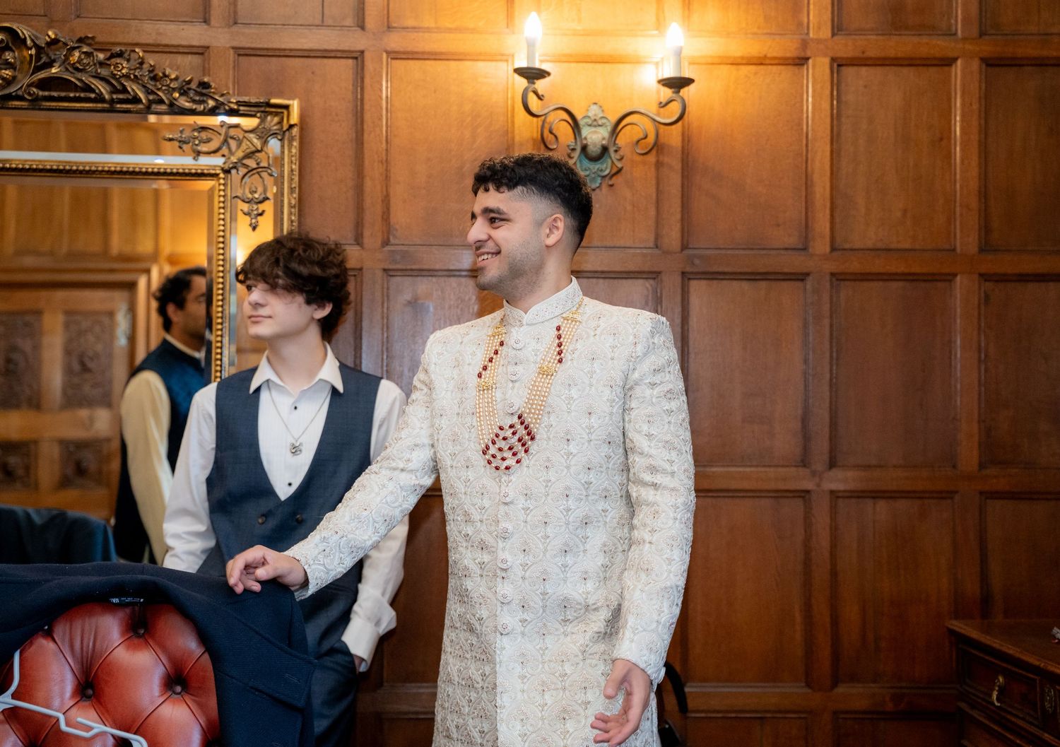 Groom in white embroidered sherwani standing with groomsman in wood-paneled wedding venue room.
