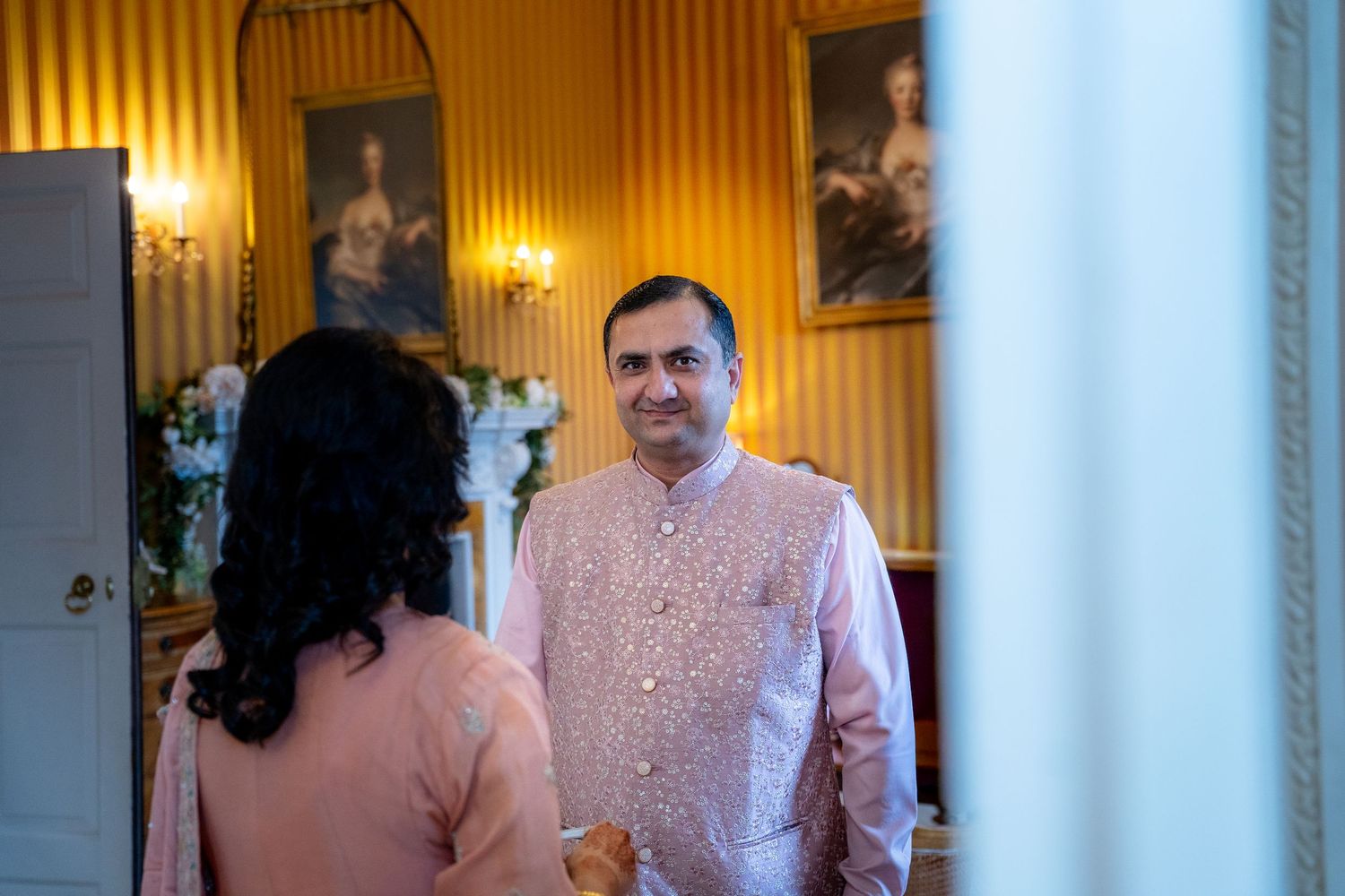 Man in pink sherwani standing in golden-walled room with portrait paintings.