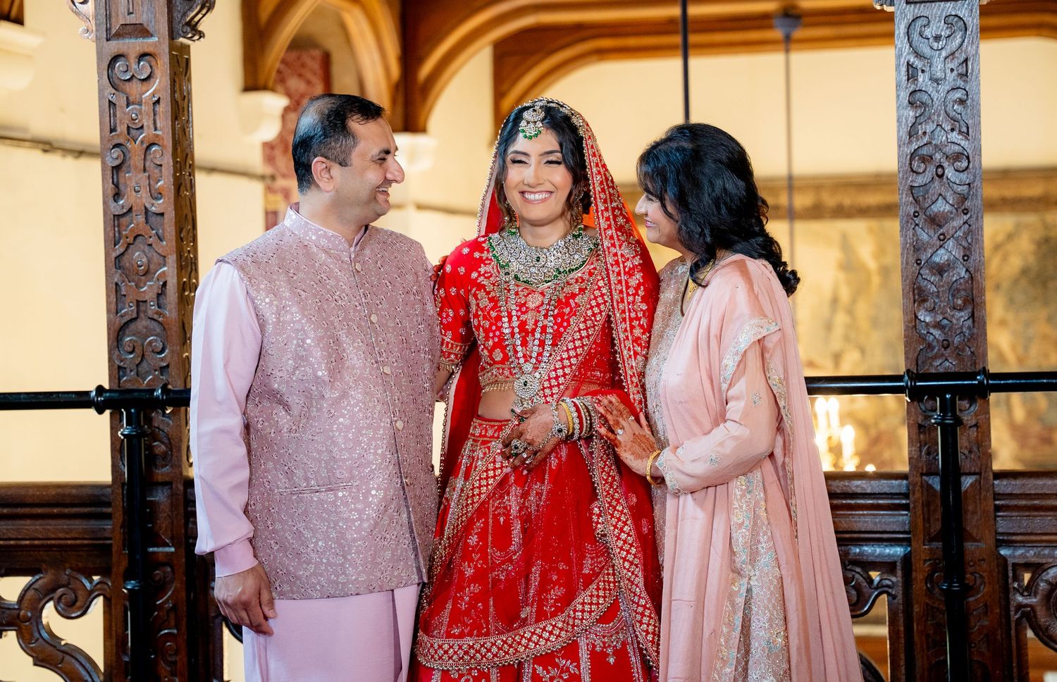 Indian bride in red bridal lehenga stands smiling with parents at traditional wedding ceremony.